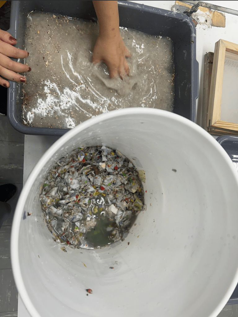 [a photo of hands swirling paper pulp around next to a bucket of torn paper scraps in water.]