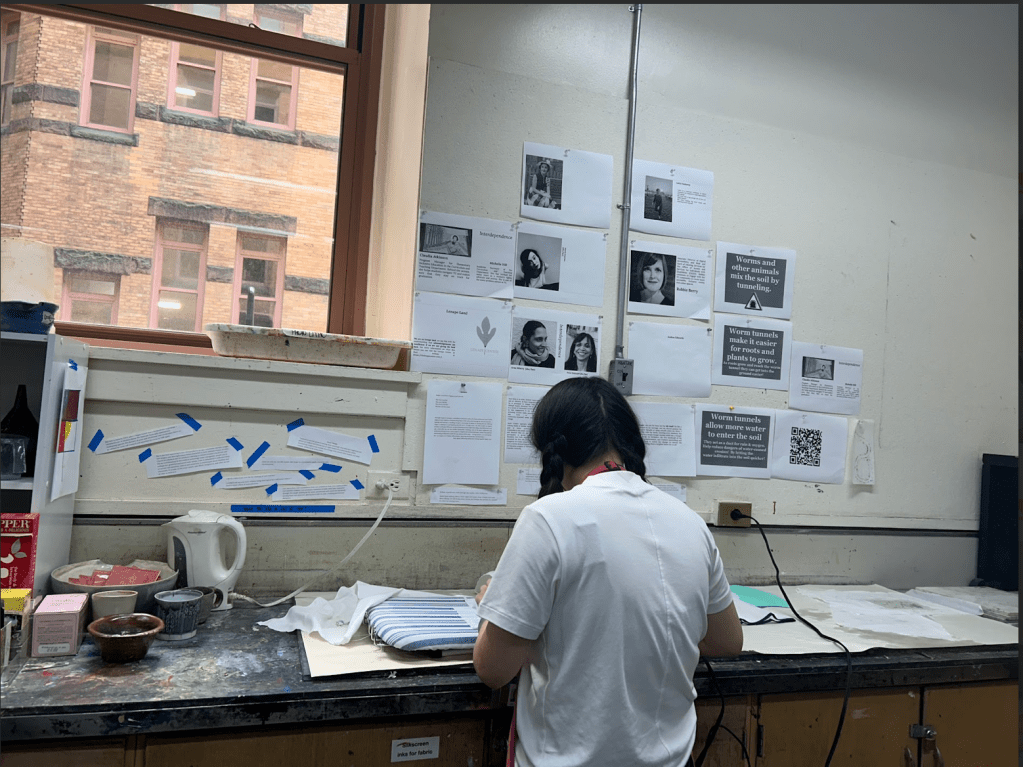 [A photo of a workshop participant working at the drying station. On the wall in front of them are various papers stuck to to it with readings, quotes, and pictures. The participant's head is bent down and working.]