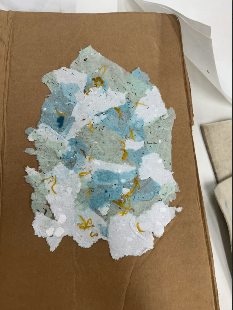 [a collage of various paper scraps neatly pressed together by a workshop participant, beginning to dry. There are blues, whites, warm yellows and many textures. The result is an artful form.]