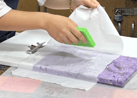 [A photo of a hand sponging a piece of wet purple paper with a lime green sponge.]