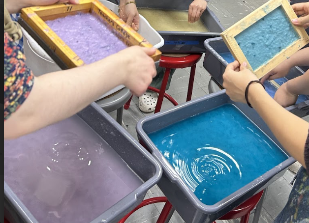 [A photo of various people working at tubs of pulp. There are hands in blue, purple, and yellow pulps.]