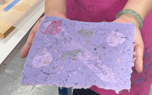 [A photo of a piece of paper created by a workshop participant. The paper is purple with fibers and materials embedded throughout. It seems thin and delicate and ephemeral.]