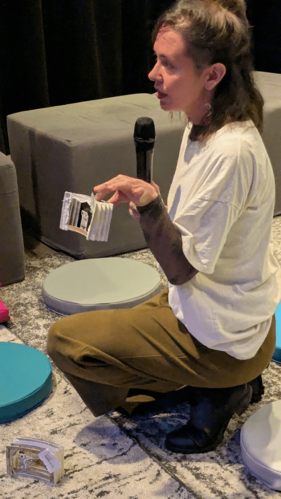 [a photo of Maria speaking into a microphone and showing two scene sketches from the play written by her, Alana, and Fred: Interference.]