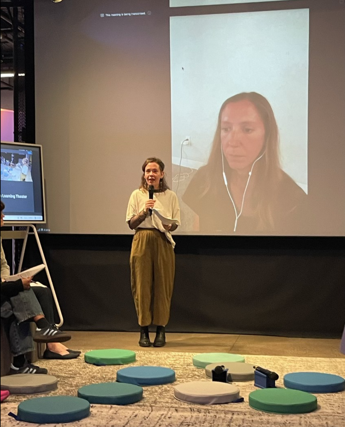 [A photo of Maria speaking into a microphone with Alana behind her on a large screen.]