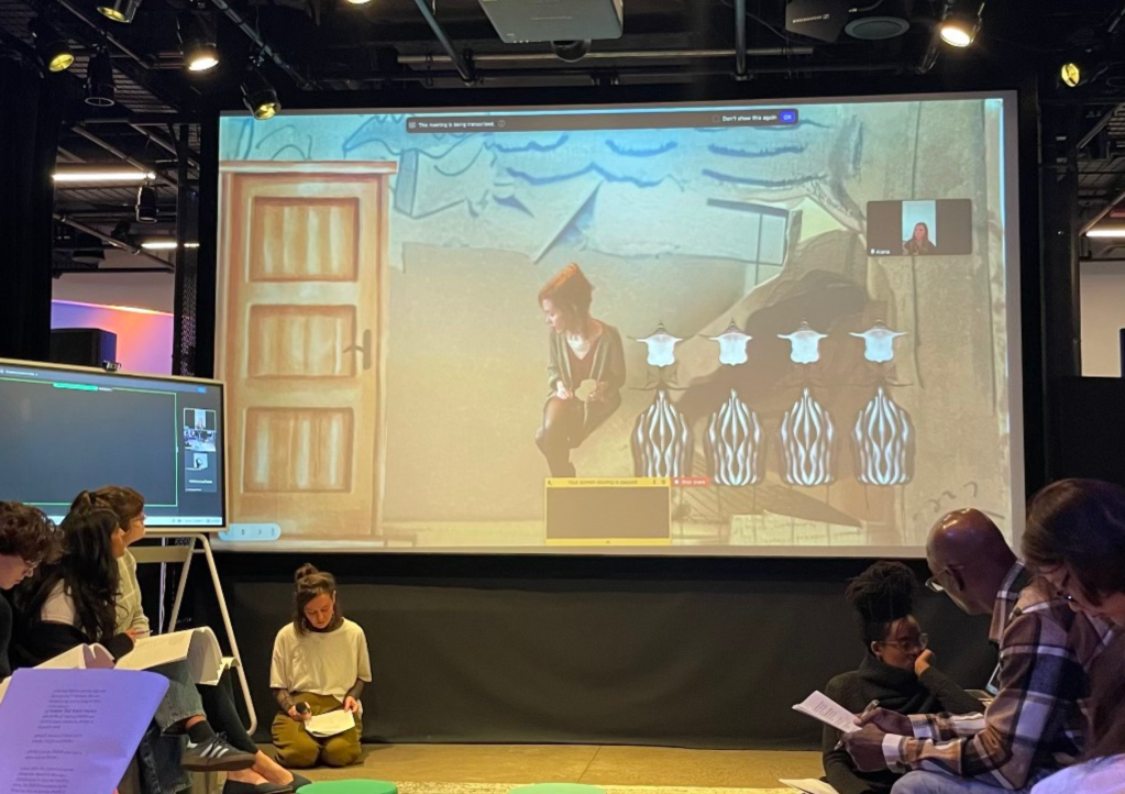 [A photo of Maria sitting on the ground with Alana projected behind her amidst various elements from Scene 2 of their play. Together they read as audience members watch and listen.]